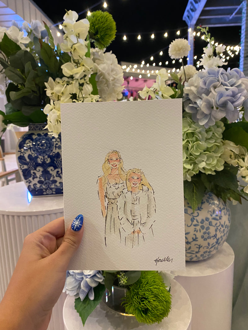 Hand holding up a small watercolor portrait illustration of a bride and a small girl. Hydrangeas and floral bouquets in the background